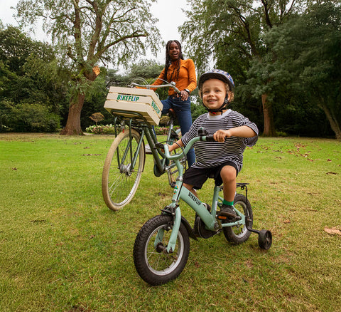 Een jongen rijdt op een groene fiets met zijwieltjes en een meisje rijdt op een groene omafiets met een mand voorop in een natuurpark met bomen en gras